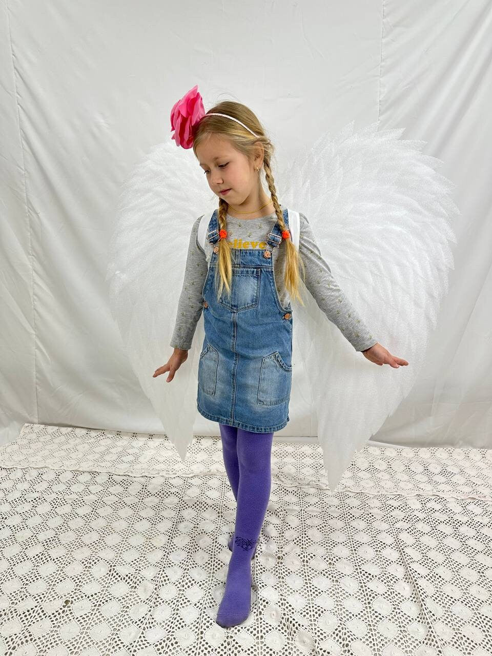 May include: A young girl wearing a denim overall dress and purple tights with white angel wings. She is wearing a pink flower headband and has her hair in two braids.