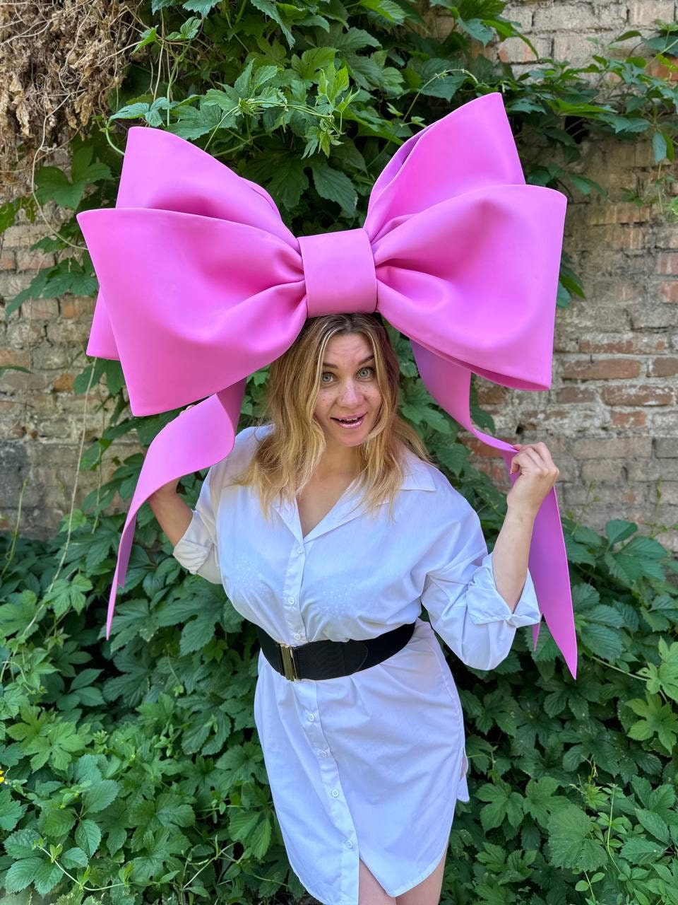May include: A large, bright pink bow, worn as a headpiece, dominates the image. The bow is made of a smooth material and is positioned atop a person's head. The person is wearing a white button-down shirt dress and a black belt. Green foliage and a brick wall form the background.
