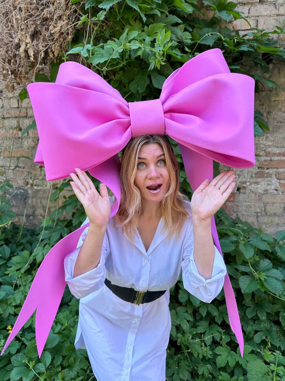 May include: A large, bright pink bow headpiece. The bow is worn on the head and has long, flowing ribbons. The person in the image is wearing a white shirt and black belt, with a green leafy background.
