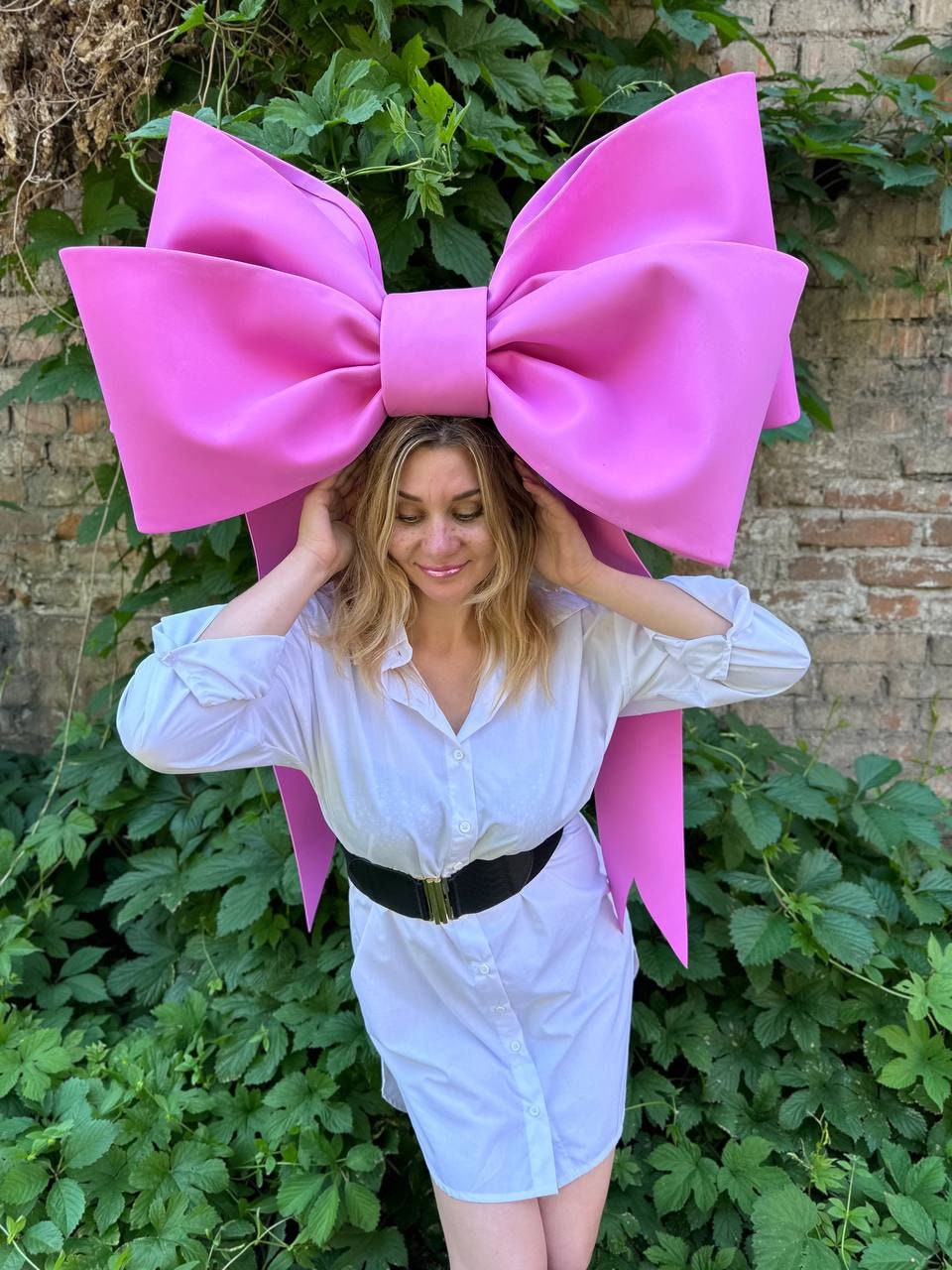 May include: A large, bright pink bow headpiece. The bow is worn on the head and is the focal point of the image. The person in the image is wearing a white button-down shirt and a black belt.