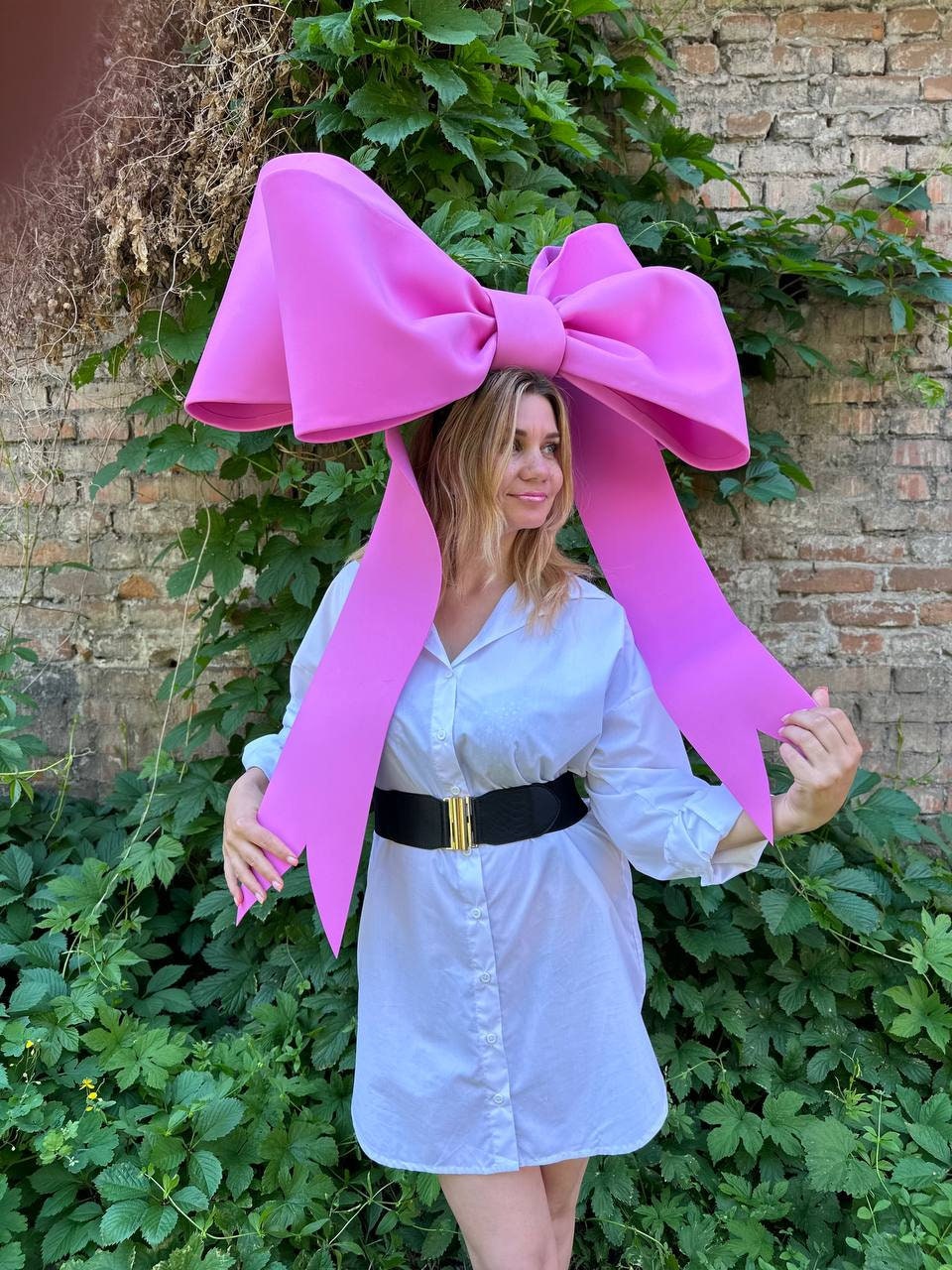 May include: A large, bright pink bow-shaped headpiece. The bow is worn on the head and has long, flowing ribbons. The person is wearing a white shirt dress with a black belt with a gold buckle. The background includes greenery and a brick wall.