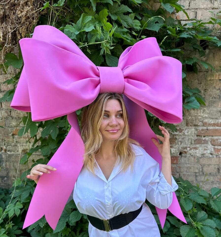 May include: A large, bright pink bow headpiece. The bow is oversized and worn on the head. The headpiece is made of a smooth material and has long, flowing ribbons. The background features green foliage and a brick wall.