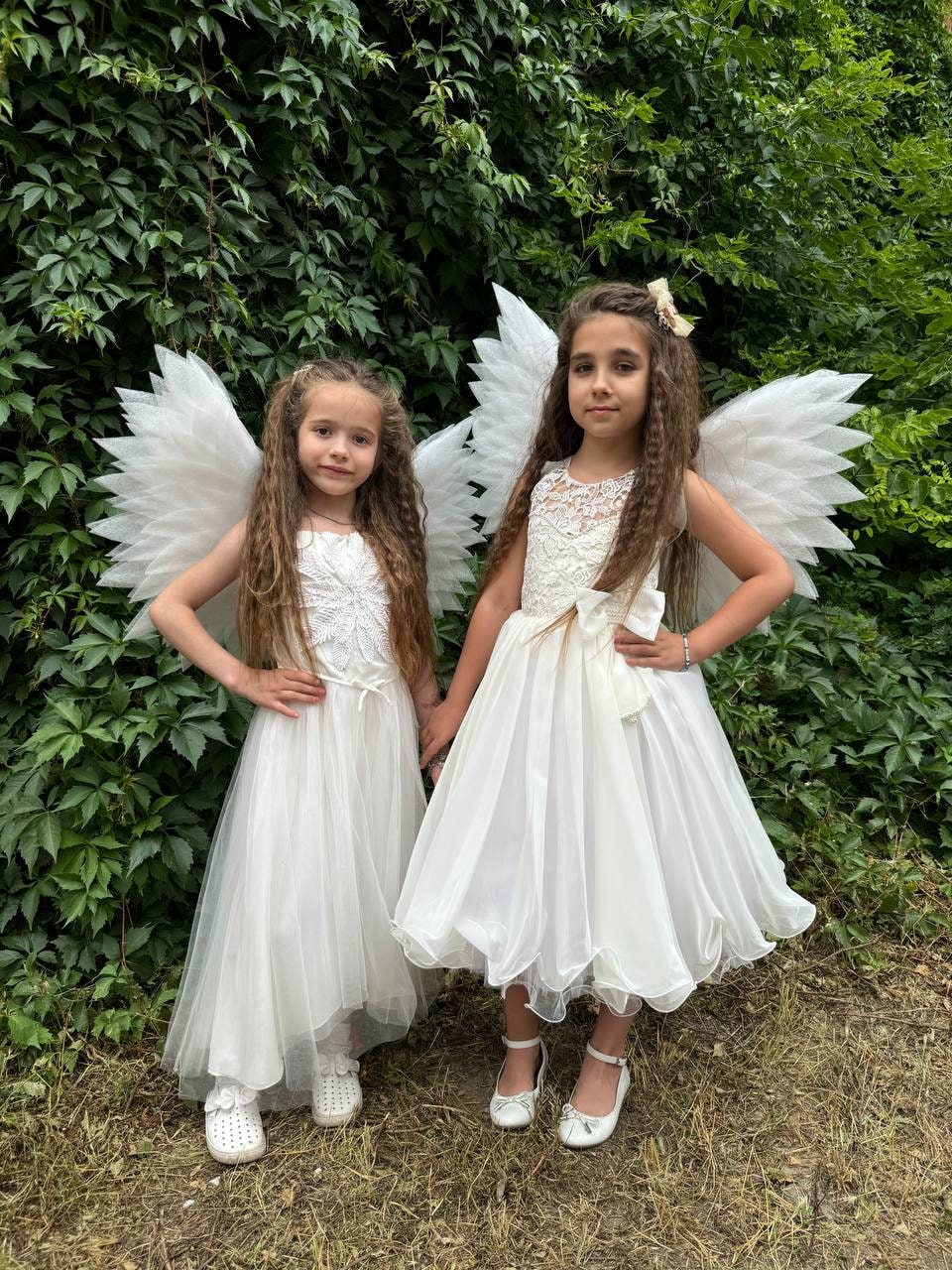 May include: Two girls wearing white angel wings and white dresses. The girls are standing in front of a green leafy background.