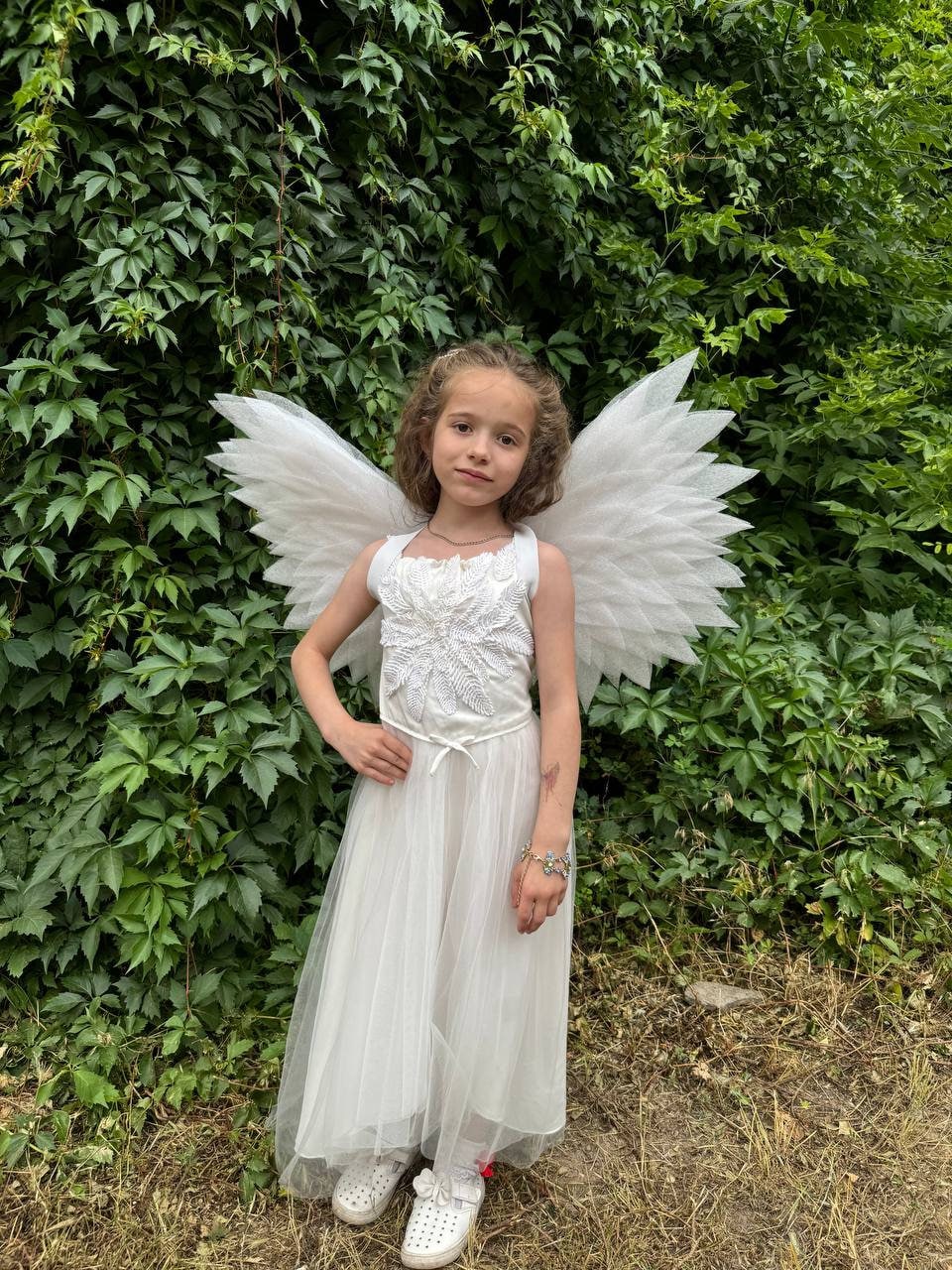 May include: A young girl wearing a white tulle dress with a white lace bodice and white angel wings. She is standing in front of a green leafy background.
