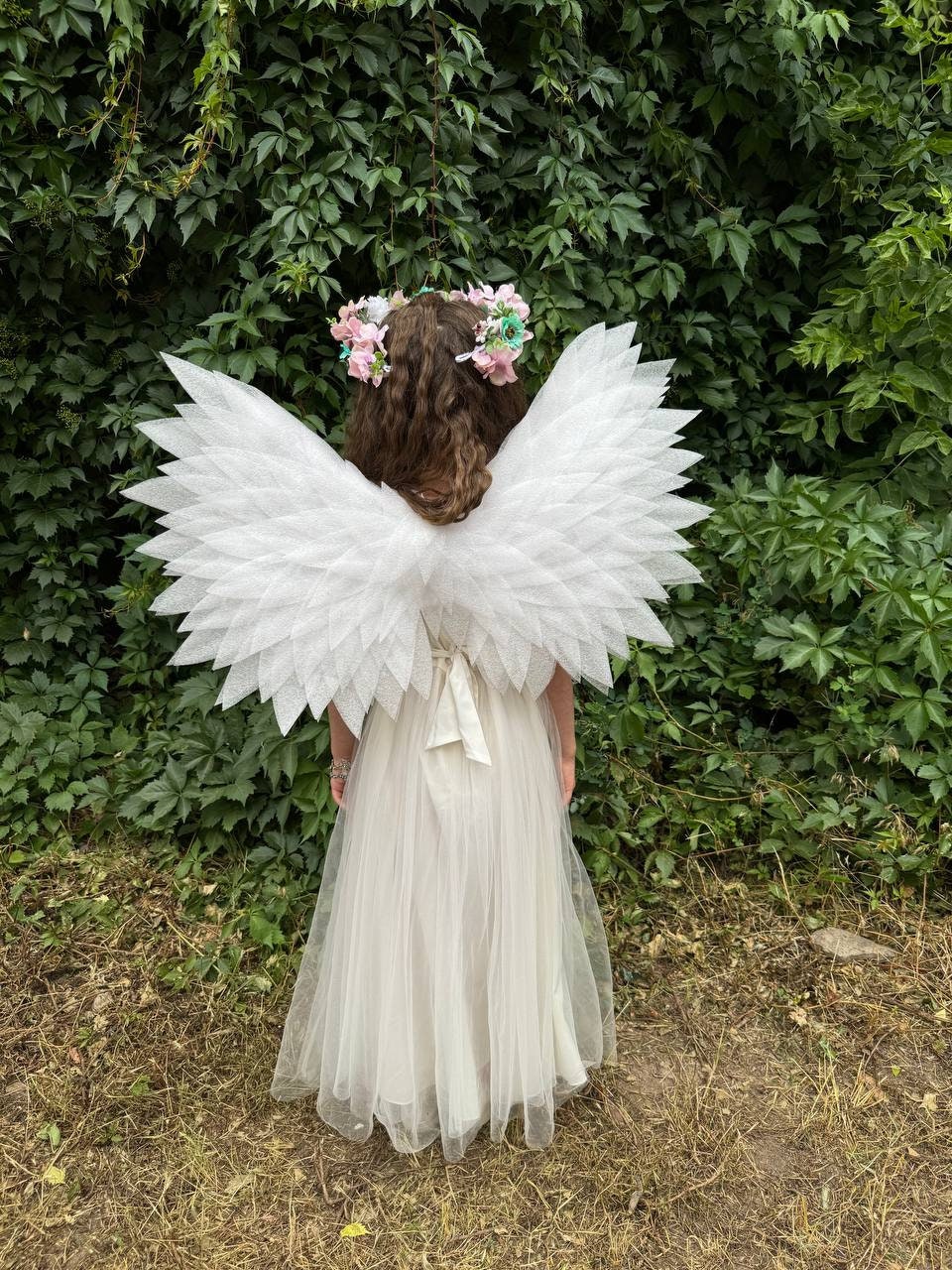 May include: A young girl wearing a white tulle dress and white angel wings with a flower crown on her head. She is standing in front of a green leafy background.