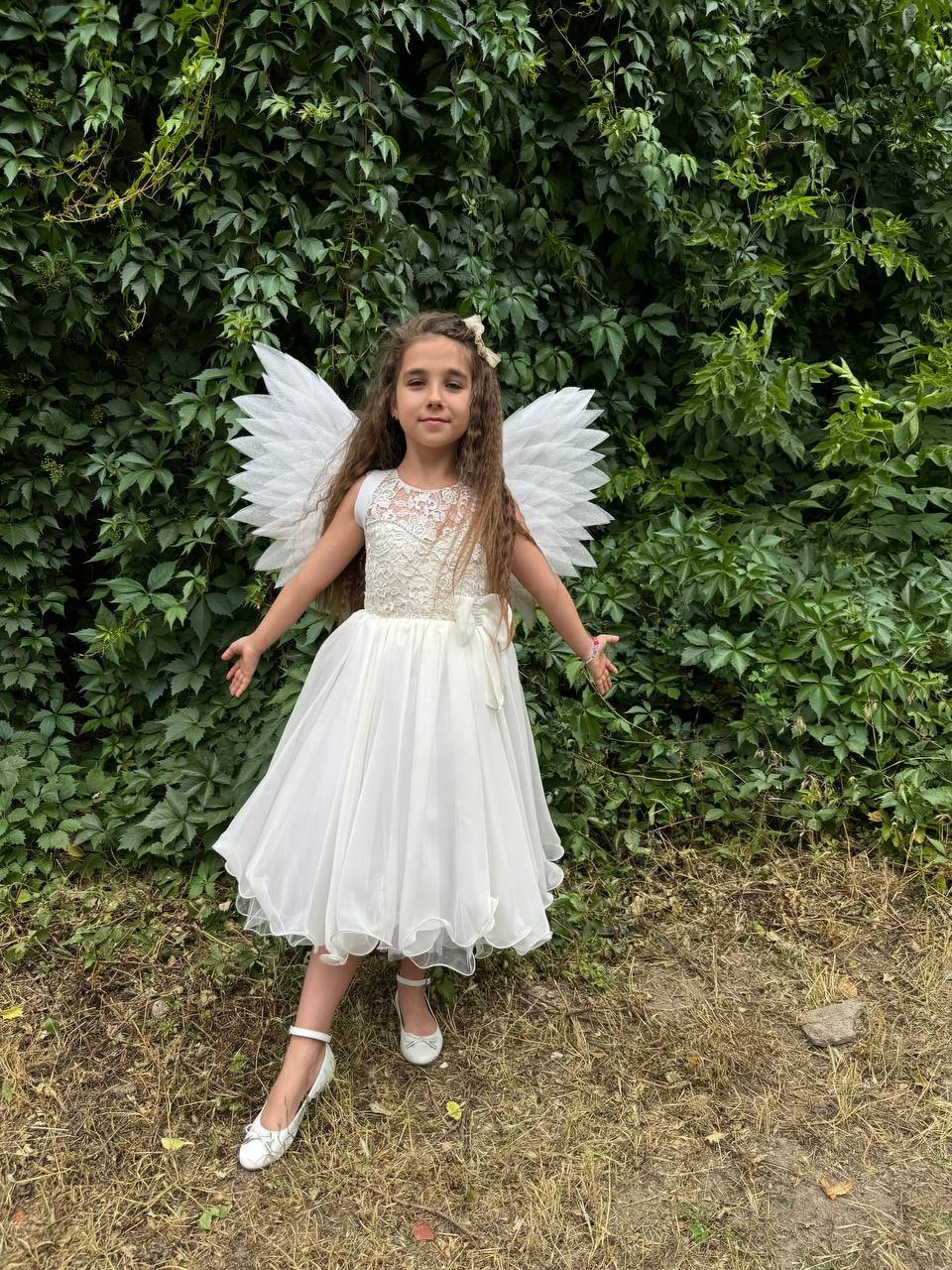 May include: A young girl wearing a white dress and angel wings, standing in front of a green leafy background.