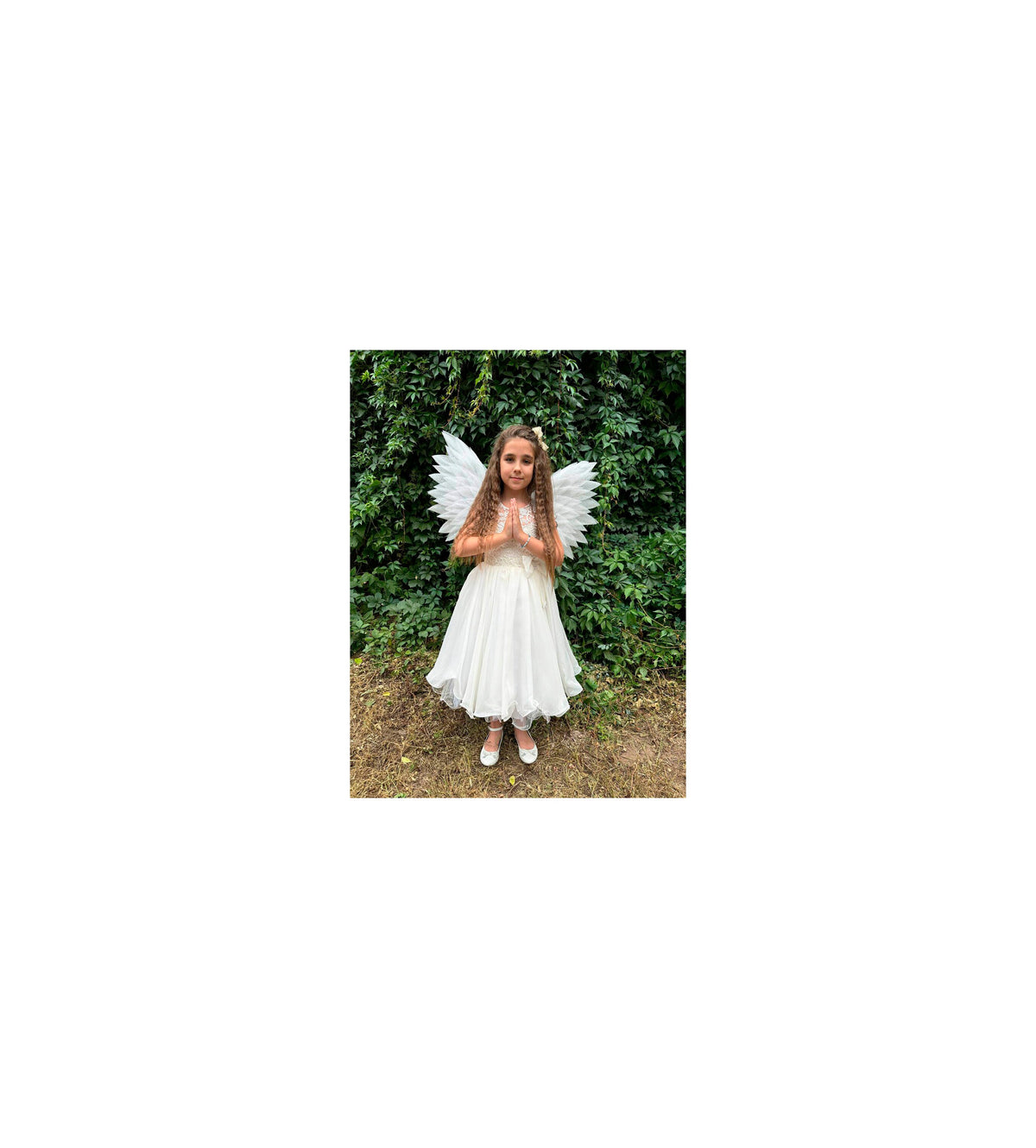 May include: A young girl wearing a white dress and white angel wings. She is standing in front of a green leafy background.