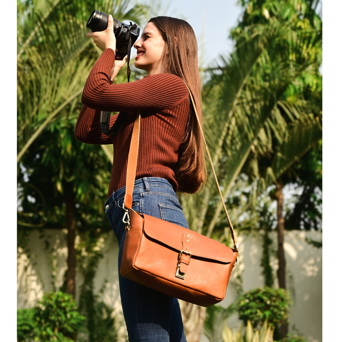 May include: A woman wearing a brown sweater and blue jeans is holding a camera and wearing a brown leather camera bag with a strap over her shoulder.