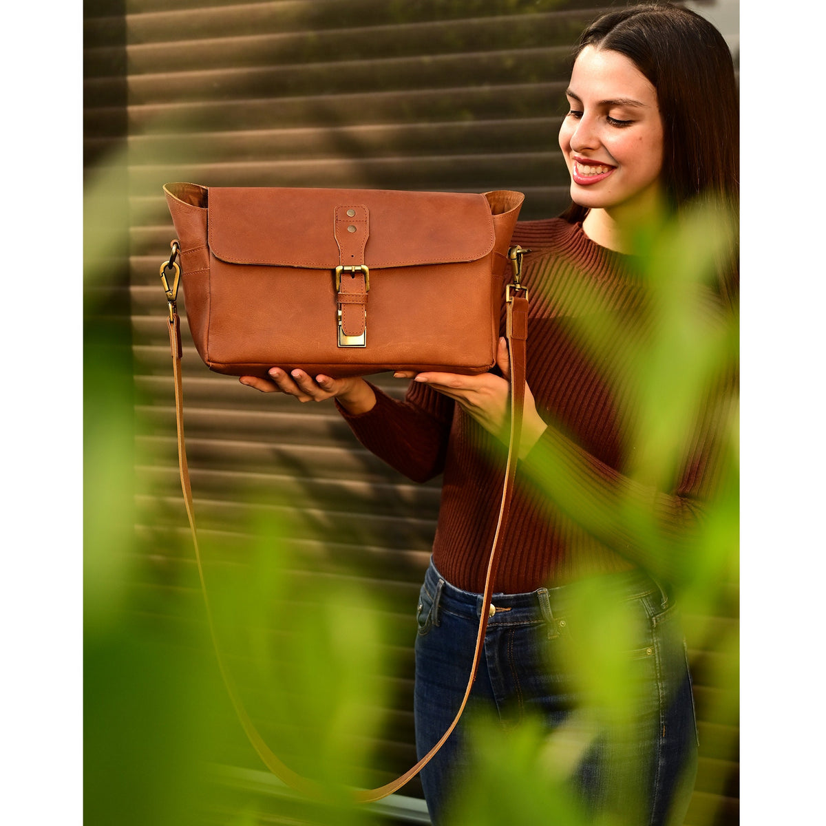 May include: A brown leather satchel bag with a flap closure and a buckle. The bag has a long adjustable strap and is being held by a person.