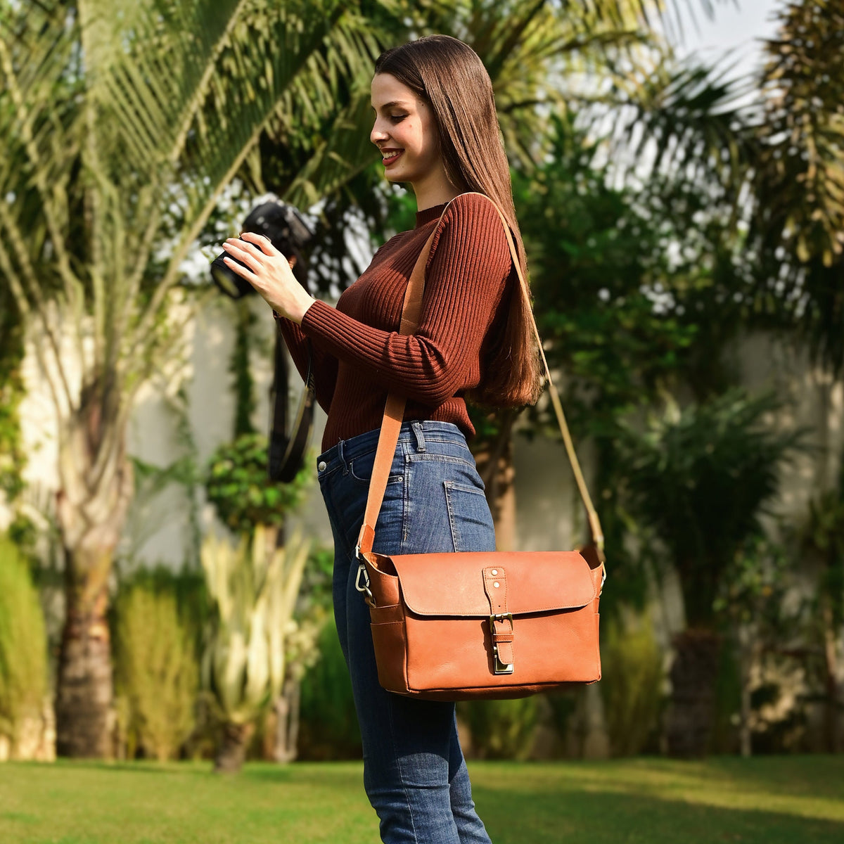 May include: A woman wearing a brown sweater and blue jeans is holding a camera and wearing a brown leather camera bag with a strap.