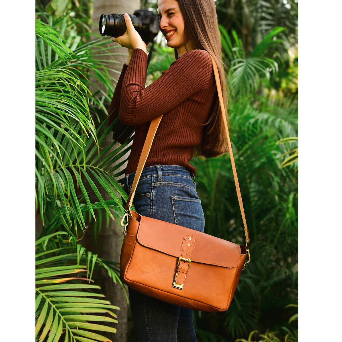 May include: A brown leather camera bag with a long adjustable strap. The bag has a flap closure with a metal buckle. The bag is worn by a person standing in front of a green leafy background.