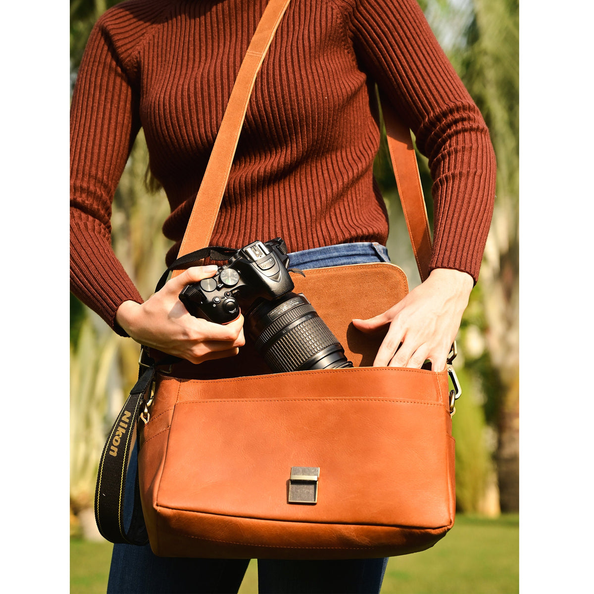 May include: A brown leather camera bag with a long adjustable strap. The bag has a flap closure and a metal buckle. A black camera with a lens is partially visible inside the bag.