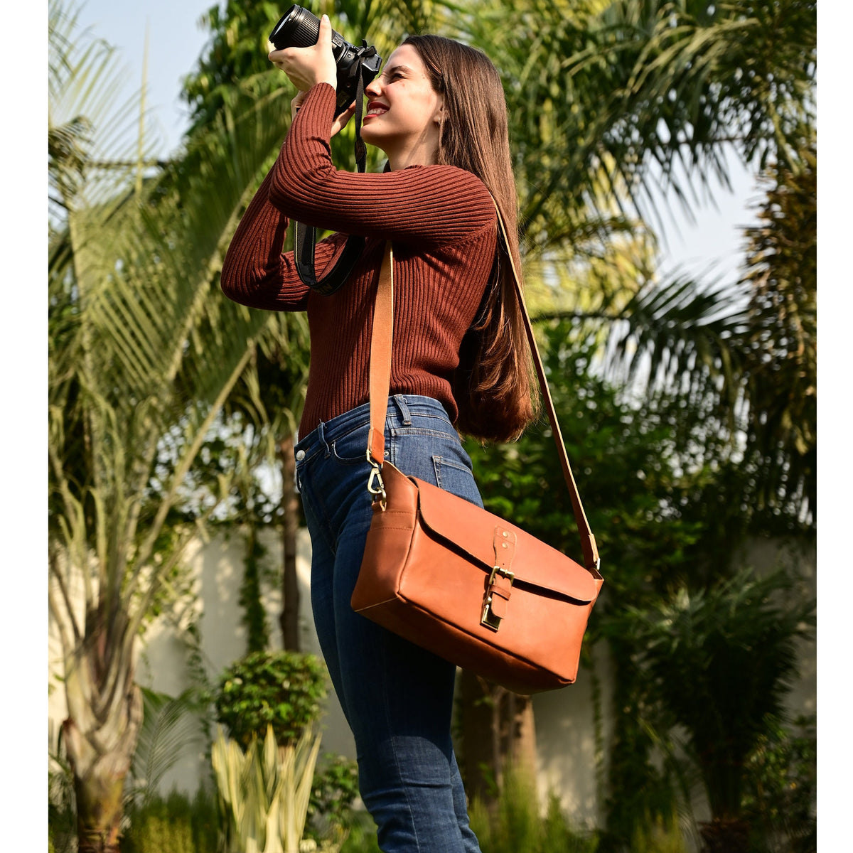May include: A woman wearing a brown sweater and blue jeans is holding a camera and wearing a brown leather messenger bag with a long strap.