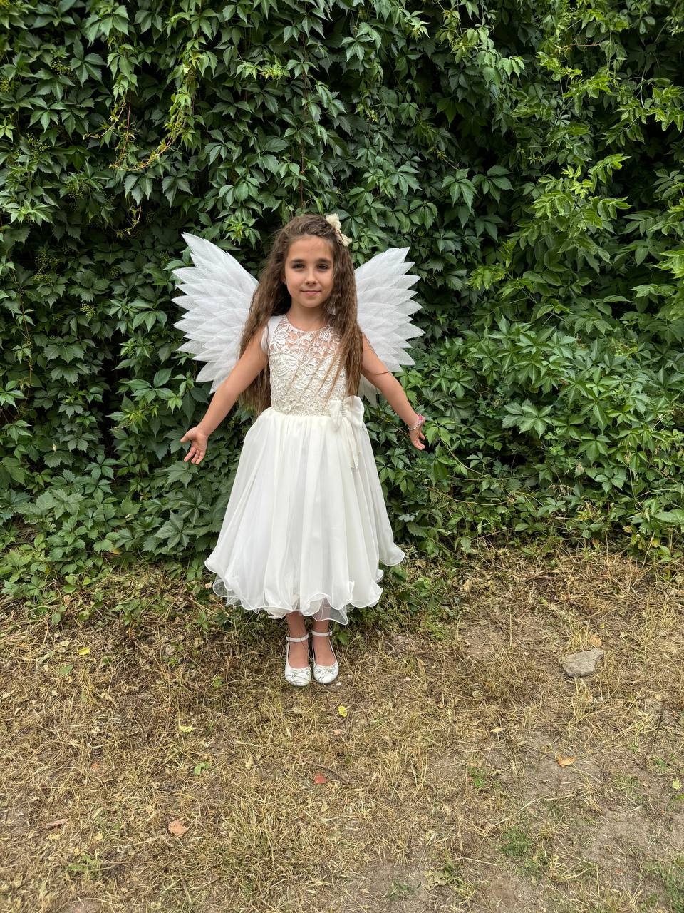 May include: A young girl wearing a white dress and white angel wings stands in front of a green leafy wall. She is smiling and looking at the camera.