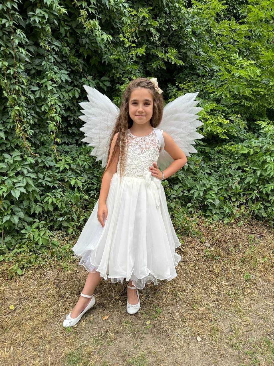 May include: A young girl wearing a white dress and white angel wings, standing in a grassy area with green foliage in the background.