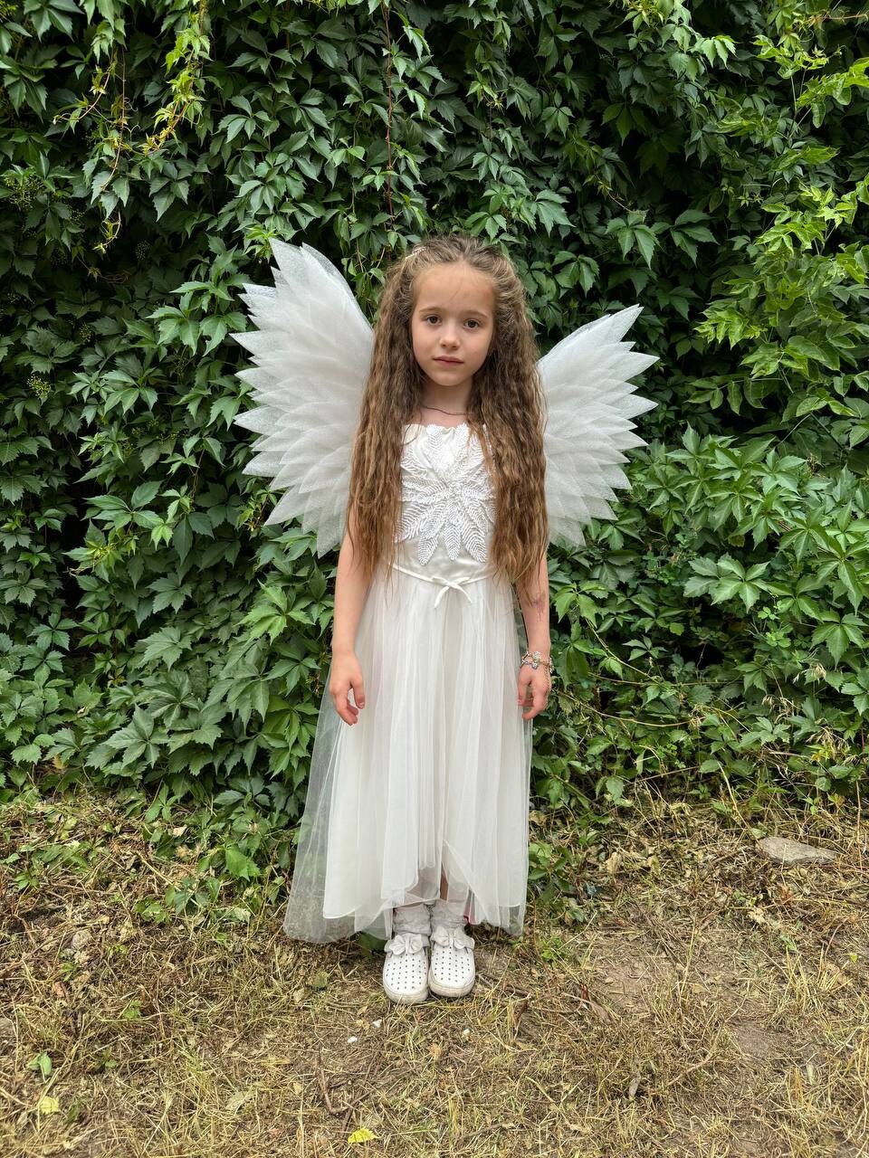 May include: A young girl wearing a white dress and angel wings. She has long brown hair and is standing in front of a green leafy background.