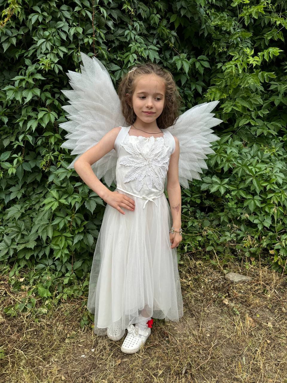 May include: A young girl wearing a white angel costume with feathery wings. She is standing in front of a green leafy background.