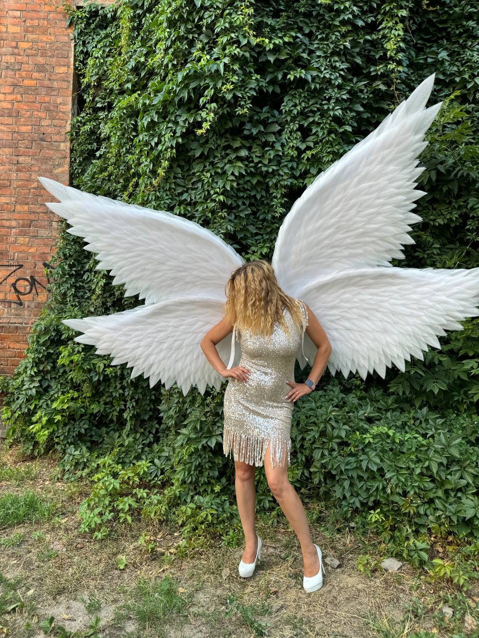 May include: A woman wearing large white angel wings and a silver sequined dress with fringe. She is also wearing white high heels. The background features a wall of green ivy and a brick wall.