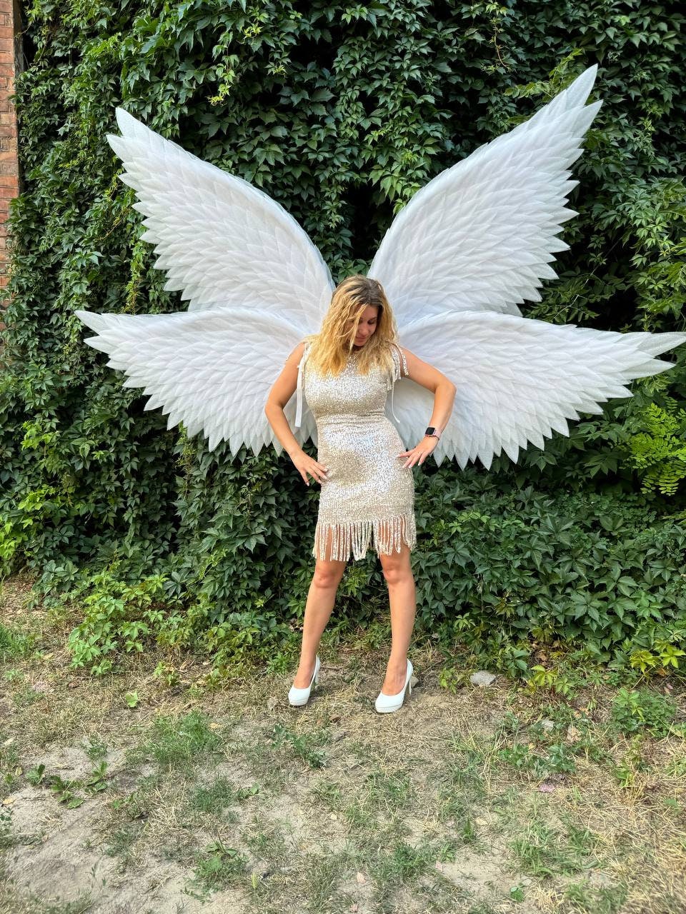 May include: A woman wearing a sparkly gold dress with fringe and white heels is standing in front of a green leafy wall. She is wearing large white angel wings.
