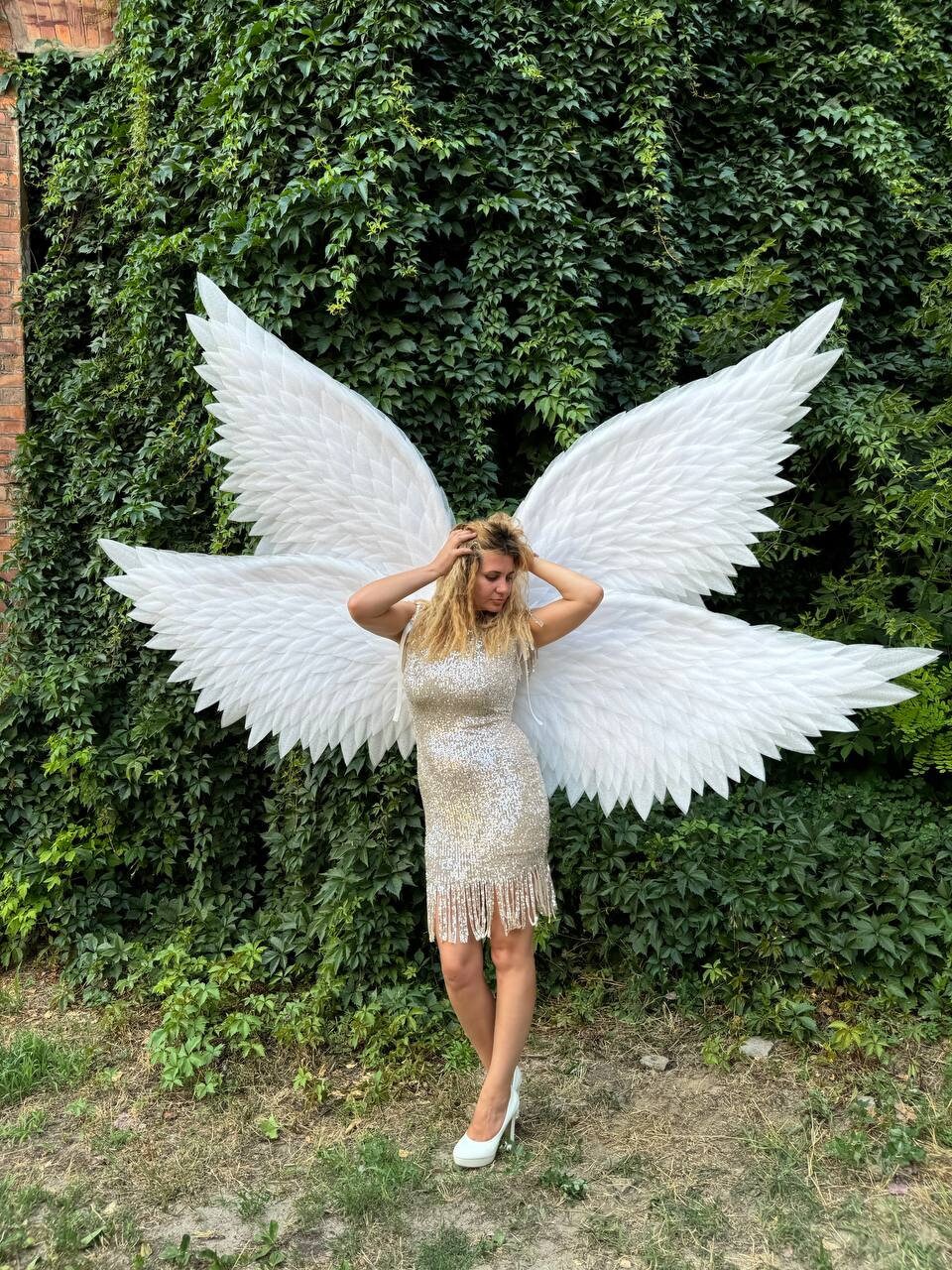 May include: A woman wearing a gold sequined dress with fringe and white high heels stands in front of a wall of green foliage. Large white angel wings are behind her, with a brick wall visible on the left.