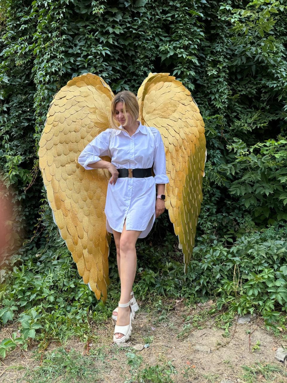 May include: A person wearing a white shirt and a black belt stands in front of a green leafy background. The person is wearing large, gold angel wings.