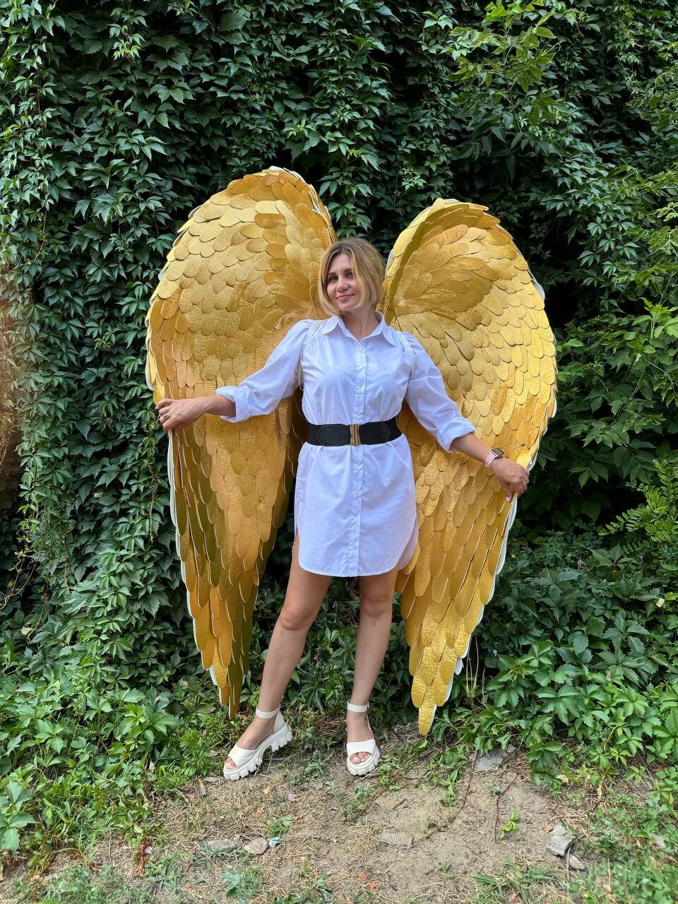 May include: A person wearing a white shirt and a black belt is standing in front of a green leafy background. They are holding large, gold angel wings.