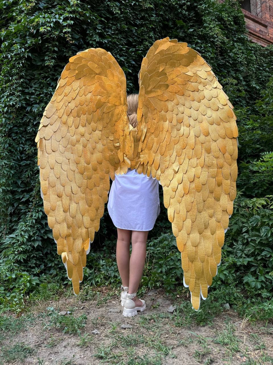 May include: A pair of large, golden angel wings with a feathery texture. The wings are made of paper or cardboard and are designed for costume or theatrical use.