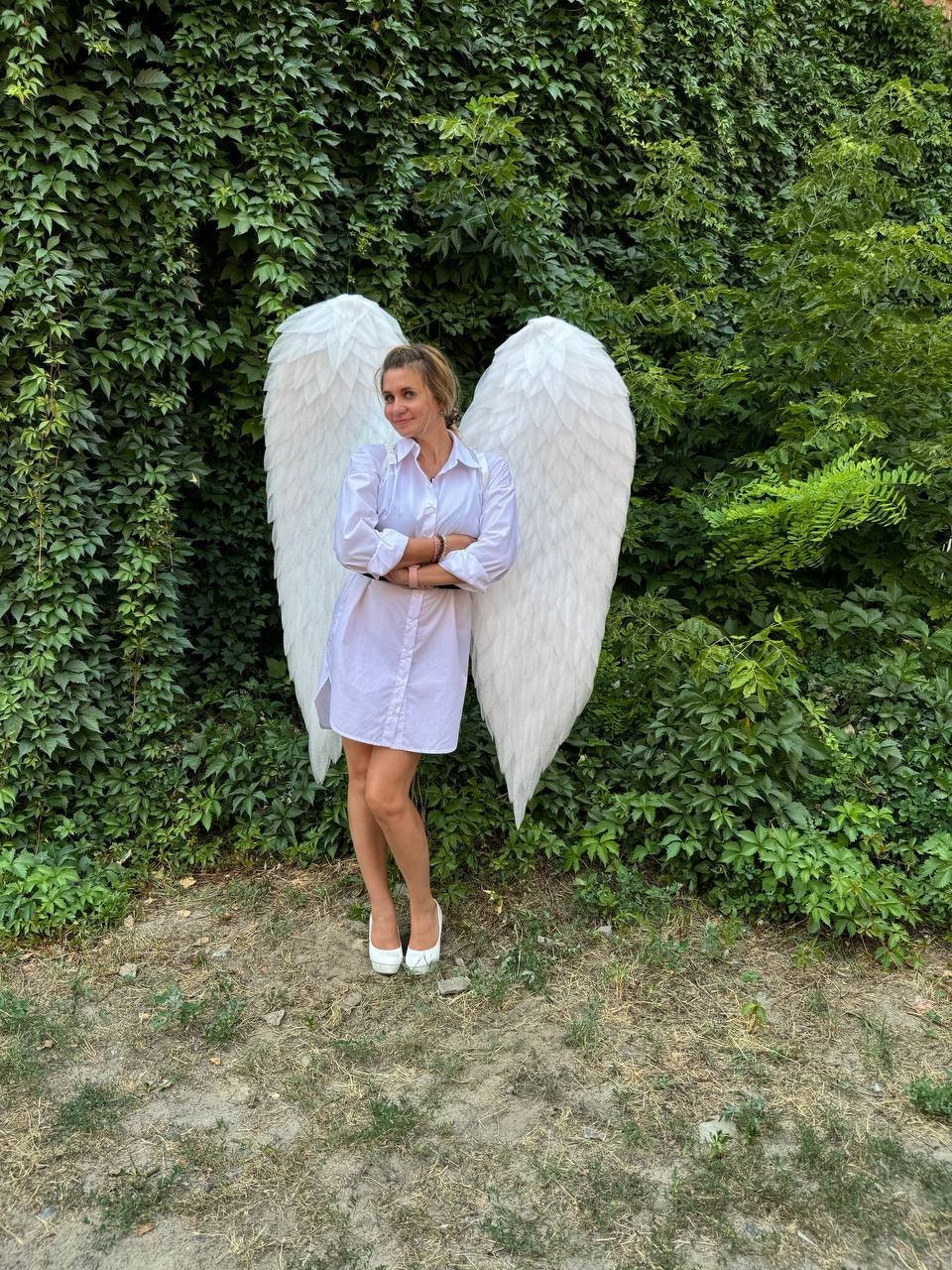 May include: A person wearing a white shirt dress and white heels stands in front of a wall of green foliage, with large white angel wings. The wings are detailed with layered feathers.