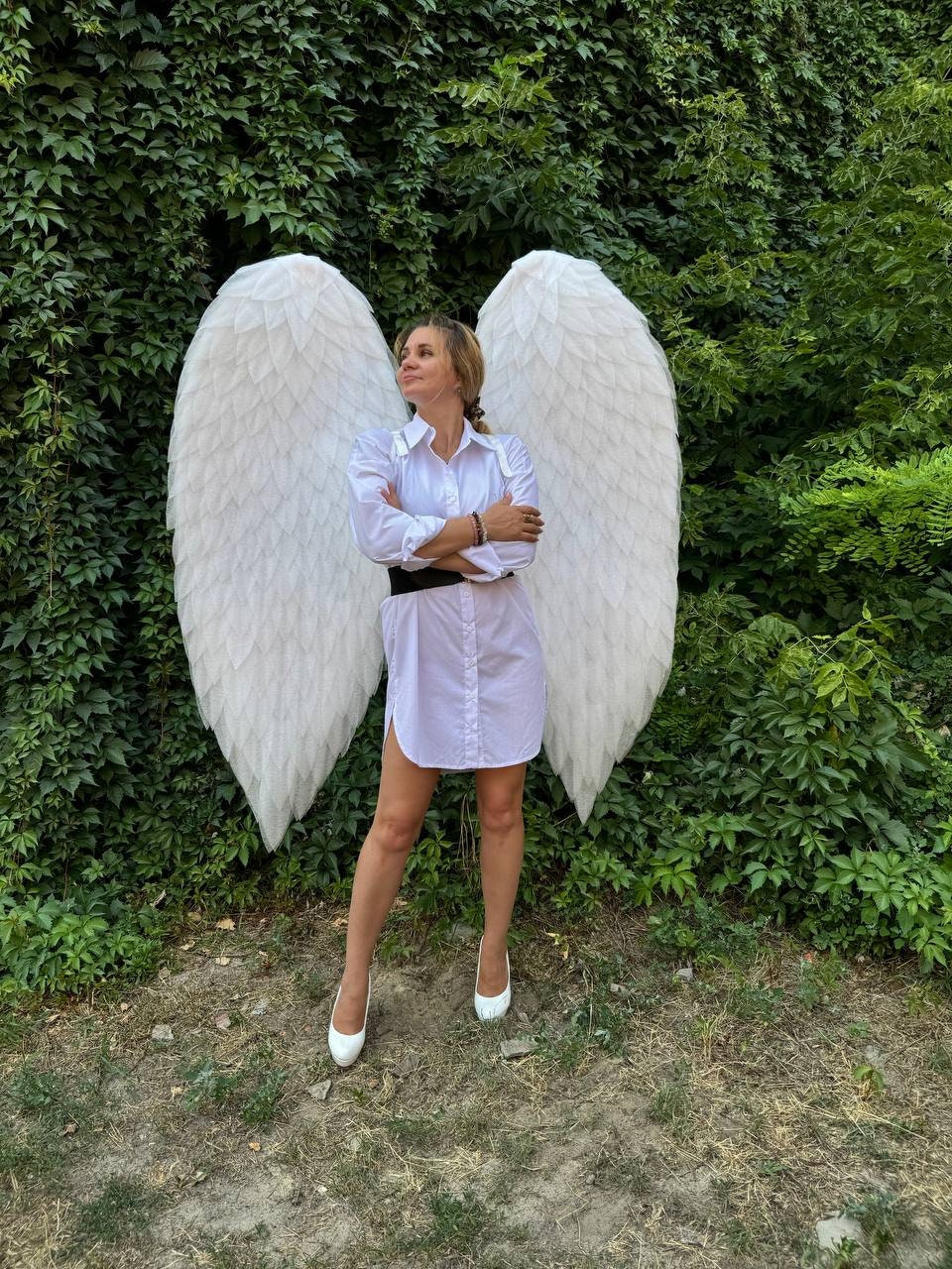 May include: A woman wearing a white shirt dress, black belt, and white heels stands in front of large white angel wings. The wings are made of feathers and are attached to a green leafy background.
