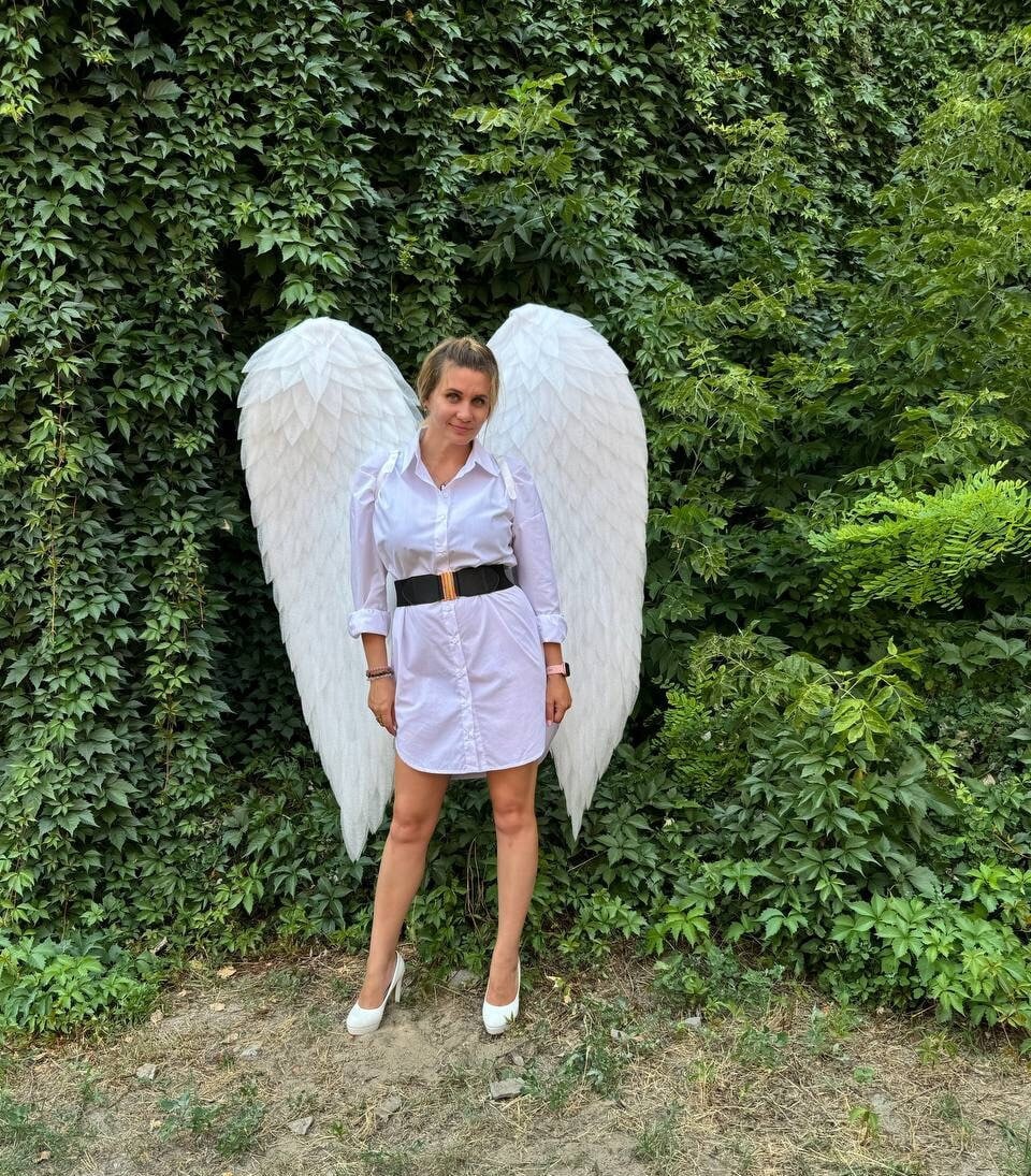May include: A white dress shirt with a black belt and white high heels. Large white angel wings are behind the person. The background is a wall of green foliage.