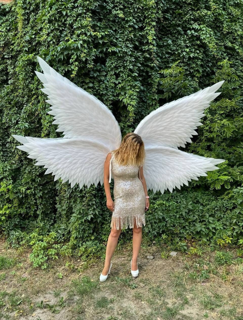 May include: A person wearing a gold sequined dress with fringe and white heels, posing in front of a wall of green foliage. Large white angel wings are attached to the person's back.