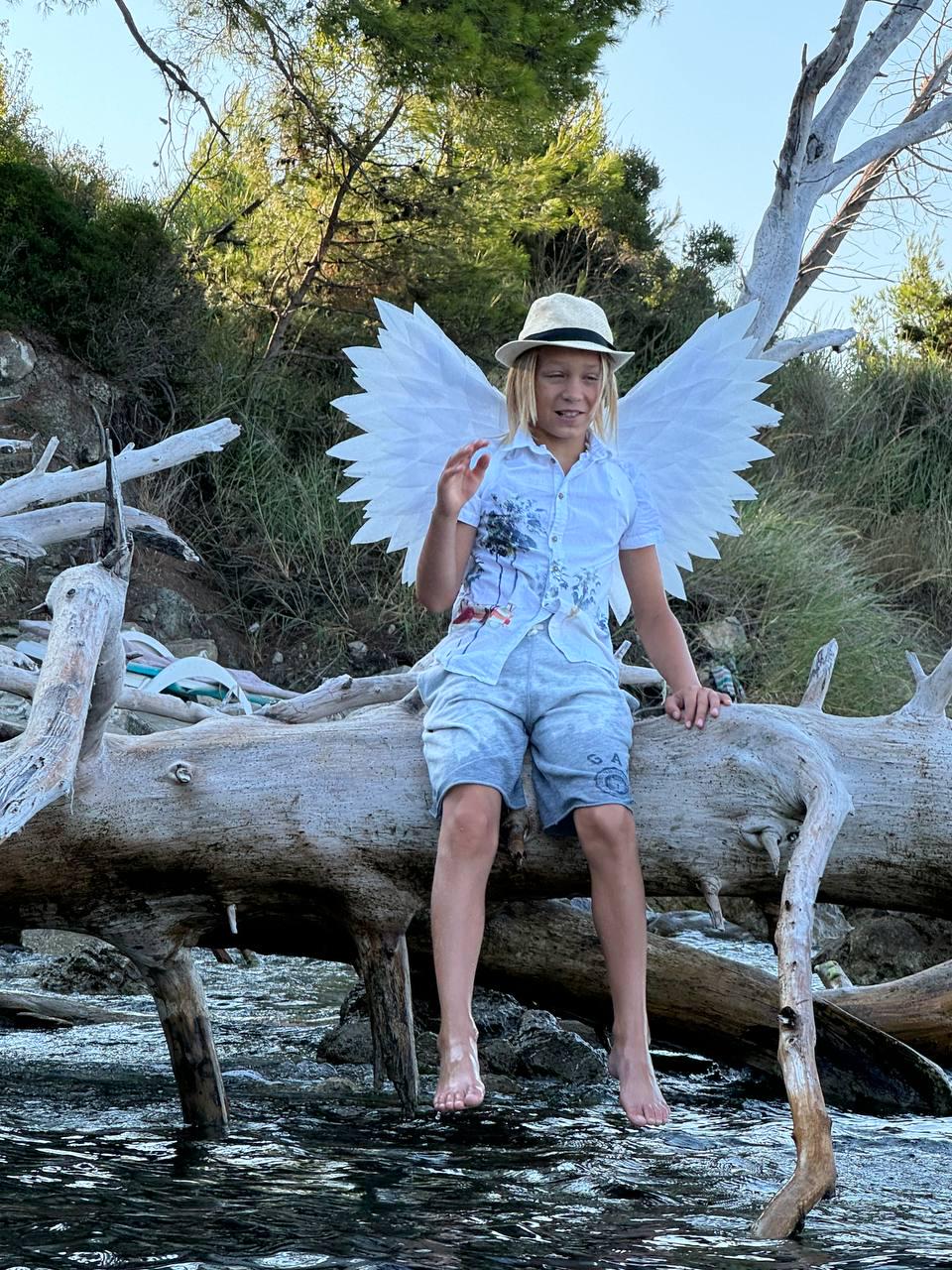May include: A young person wearing a white shirt, blue shorts, and a white hat with a white feather angel wing costume sitting on a weathered log over a stream.