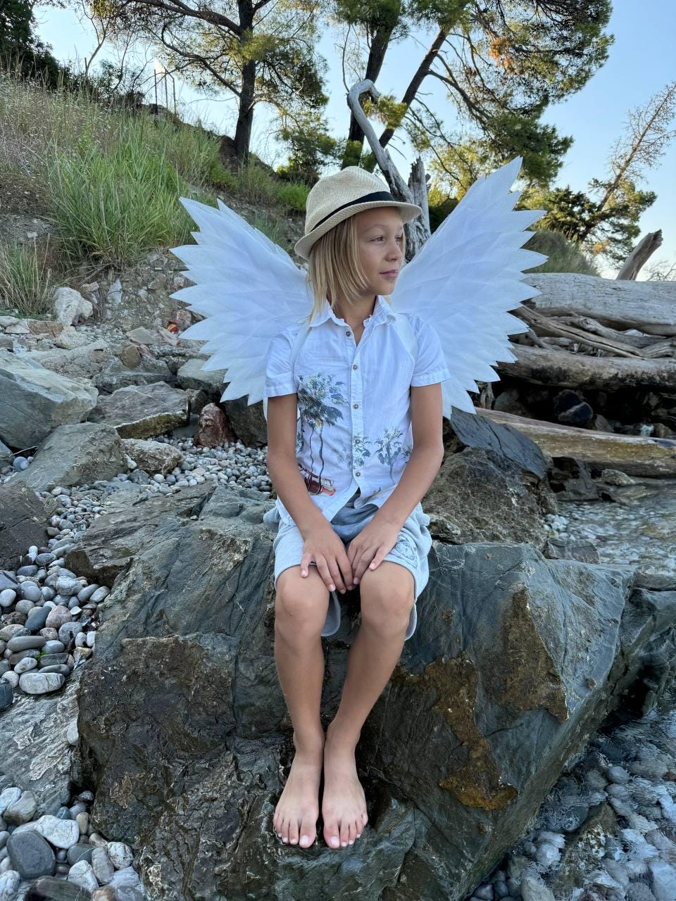 May include: A young person wearing a white shirt and a straw hat sits on a rock with large white angel wings. The person is sitting with their legs crossed and their hands in their lap. The background is a rocky beach with green foliage.