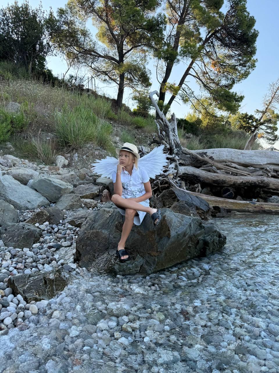 May include: A young person wearing a white dress and a straw hat sits on a rock by the water. They are wearing white angel wings and have a thoughtful expression on their face.