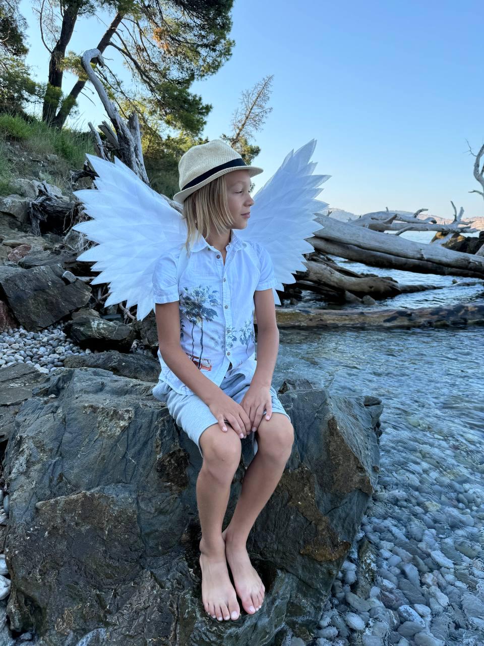 May include: A young person wearing a white shirt and a straw hat sits on a rock by the water. They are wearing large white angel wings.