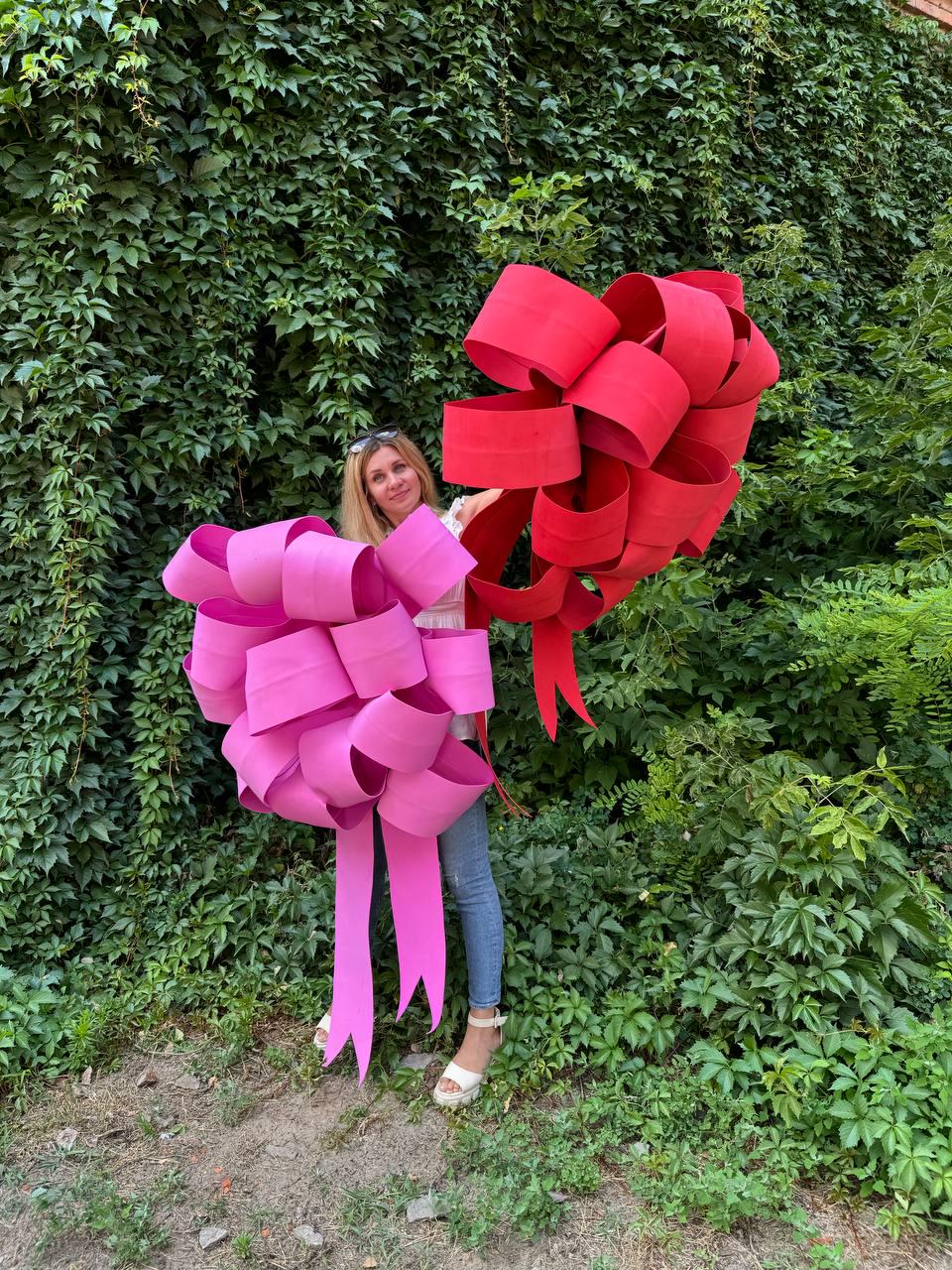 May include: A woman holds two large decorative bows. One bow is a vibrant red, and the other is a bright pink. The bows are made of thick, rolled paper or fabric, with long, flowing tails. The background is a lush green wall of foliage.