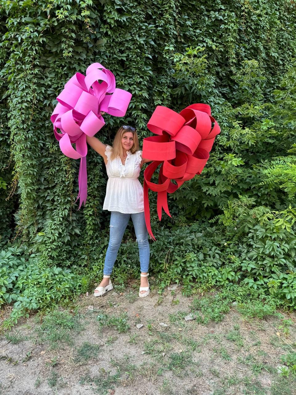 May include: A woman holding two large decorative bows. One bow is bright pink, and the other is red. The bows are made of wide, looped ribbons. The background is a wall of green foliage.