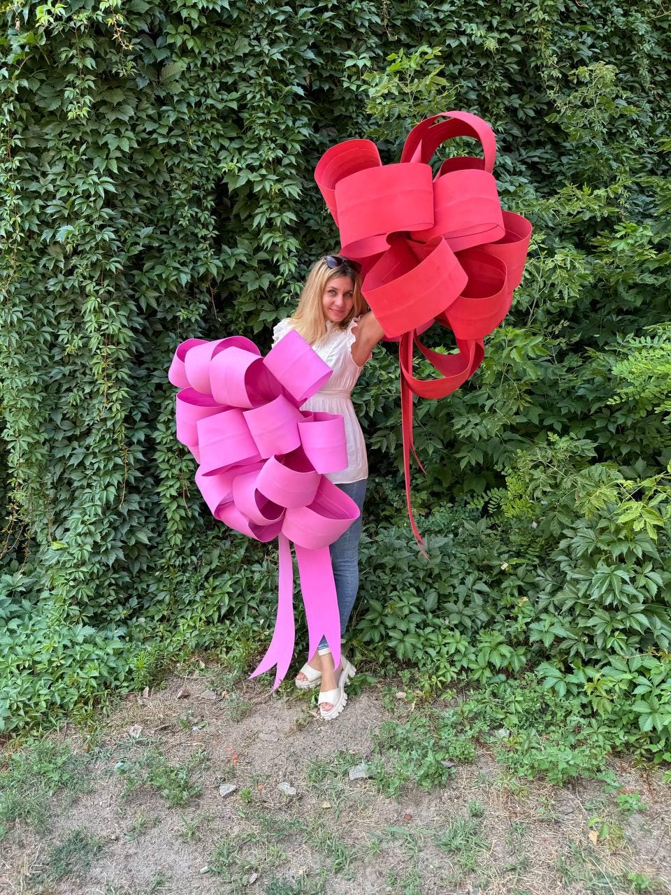 May include: A woman stands in front of a wall of green foliage, holding two large, decorative bows. One bow is bright pink, the other is red. The bows are made of wide, looped ribbons, with long, flowing tails.
