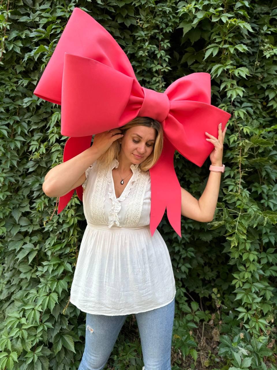 May include: A woman wearing a white top and blue jeans is standing in front of a green leafy wall. She is wearing a large, bright pink bow on her head.
