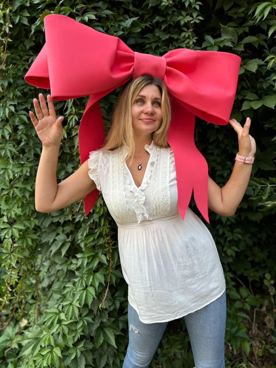 May include: A woman wearing a large, bright pink bow on her head. The bow is made of fabric and is tied in a large knot. The woman is smiling and looking at the camera. She is wearing a white top and blue jeans.