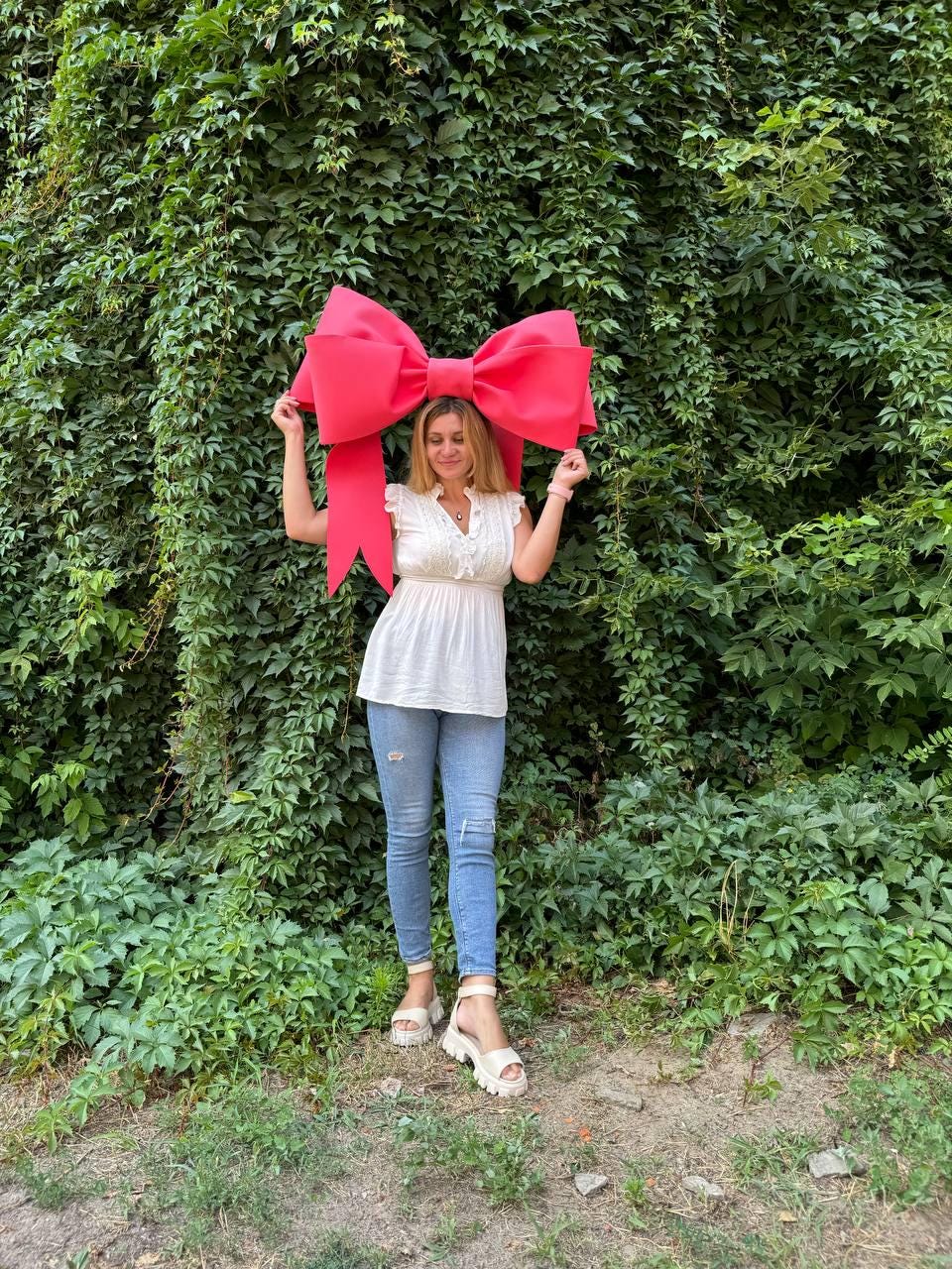 May include: A woman is holding a large, bright pink bow in front of a green ivy wall. She is wearing a white blouse, blue jeans, and white sandals.