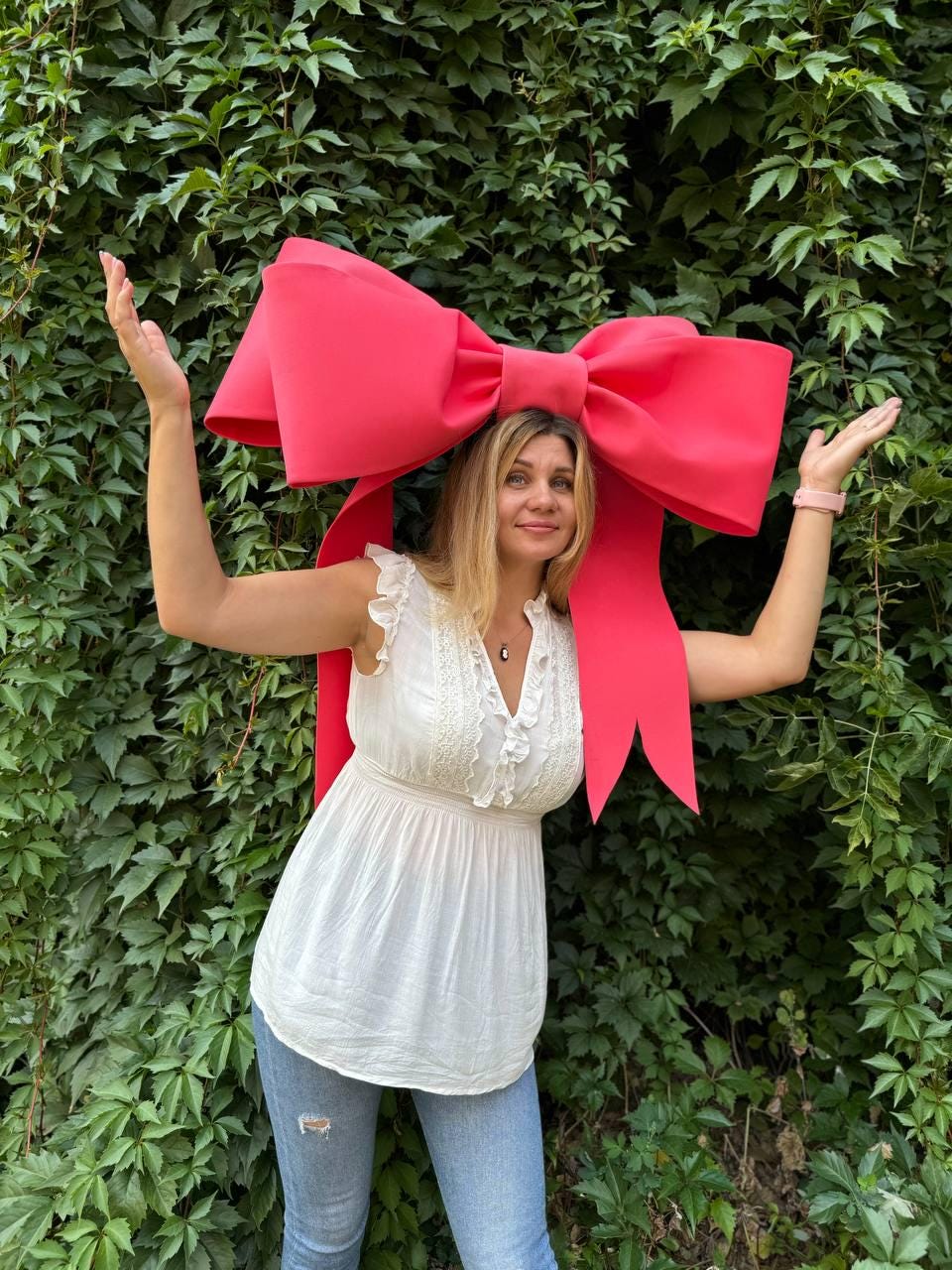 May include: A woman wearing a white top and blue jeans is standing in front of a green leafy wall. She is wearing a large, bright pink bow on her head.