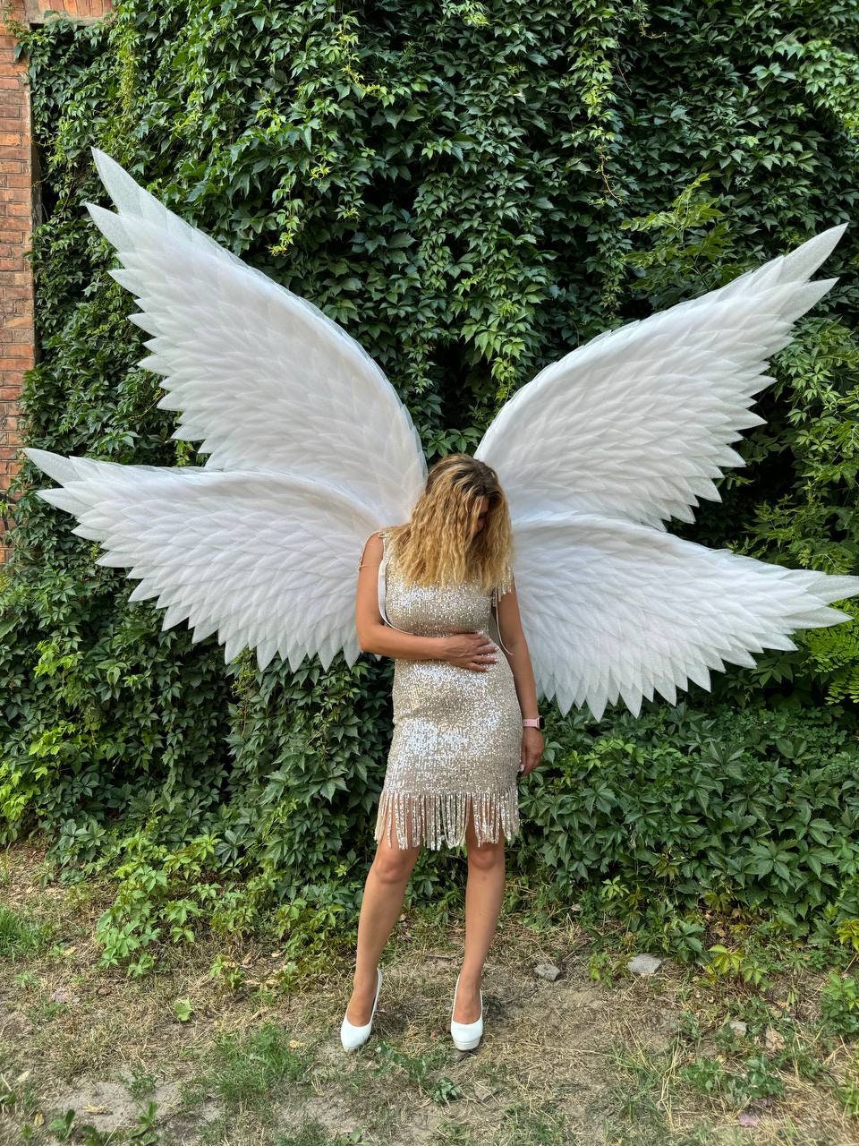 May include: A person wearing a sparkly gold dress with fringe and white heels stands in front of a green leafy wall with large white angel wings.