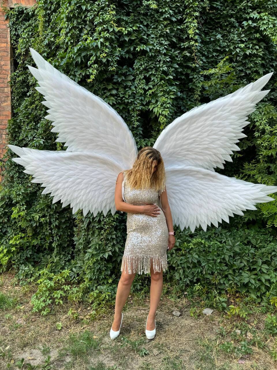 May include: A person wearing large, white angel wings and a gold sequined dress with fringe. The wings are made of white feathers and are spread wide. The person is wearing white high heels. The background is a wall of green foliage.