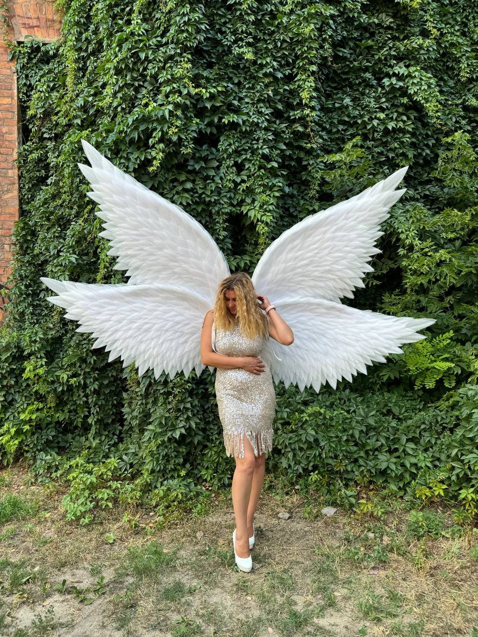 May include: A person wearing large white angel wings and a sequined dress stands in front of a wall of green foliage. The wings are made of white feathers and are spread wide. The dress is gold and has fringe.