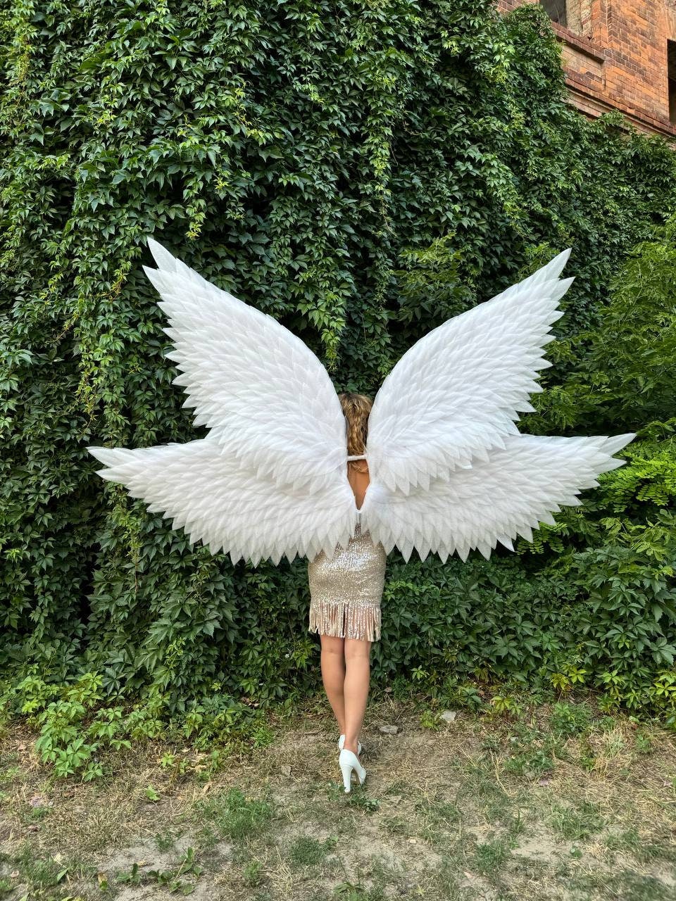 May include: A person wearing a sparkly gold dress and white angel wings with a green ivy wall in the background.