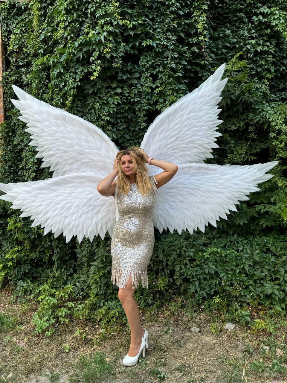 May include: A woman wearing a gold sequined dress with fringe and white high heels stands in front of large white angel wings. The wings are made of feathers and are spread out behind her. The background is a wall of green foliage.