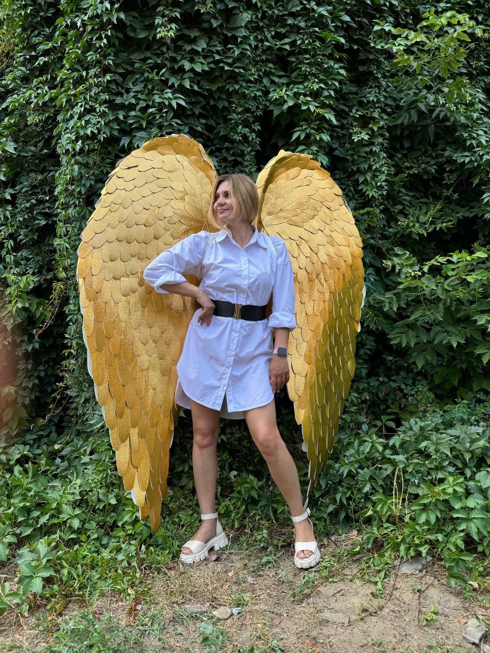 May include: A person wearing a white shirt and white sandals stands in front of a green leafy background. The person is holding large, gold, feathered angel wings.