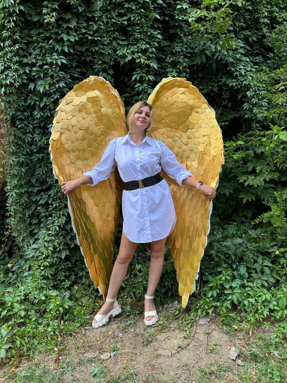 May include: A woman wearing a white shirt and a black belt is standing in front of a green leafy background. She is holding large, gold angel wings.