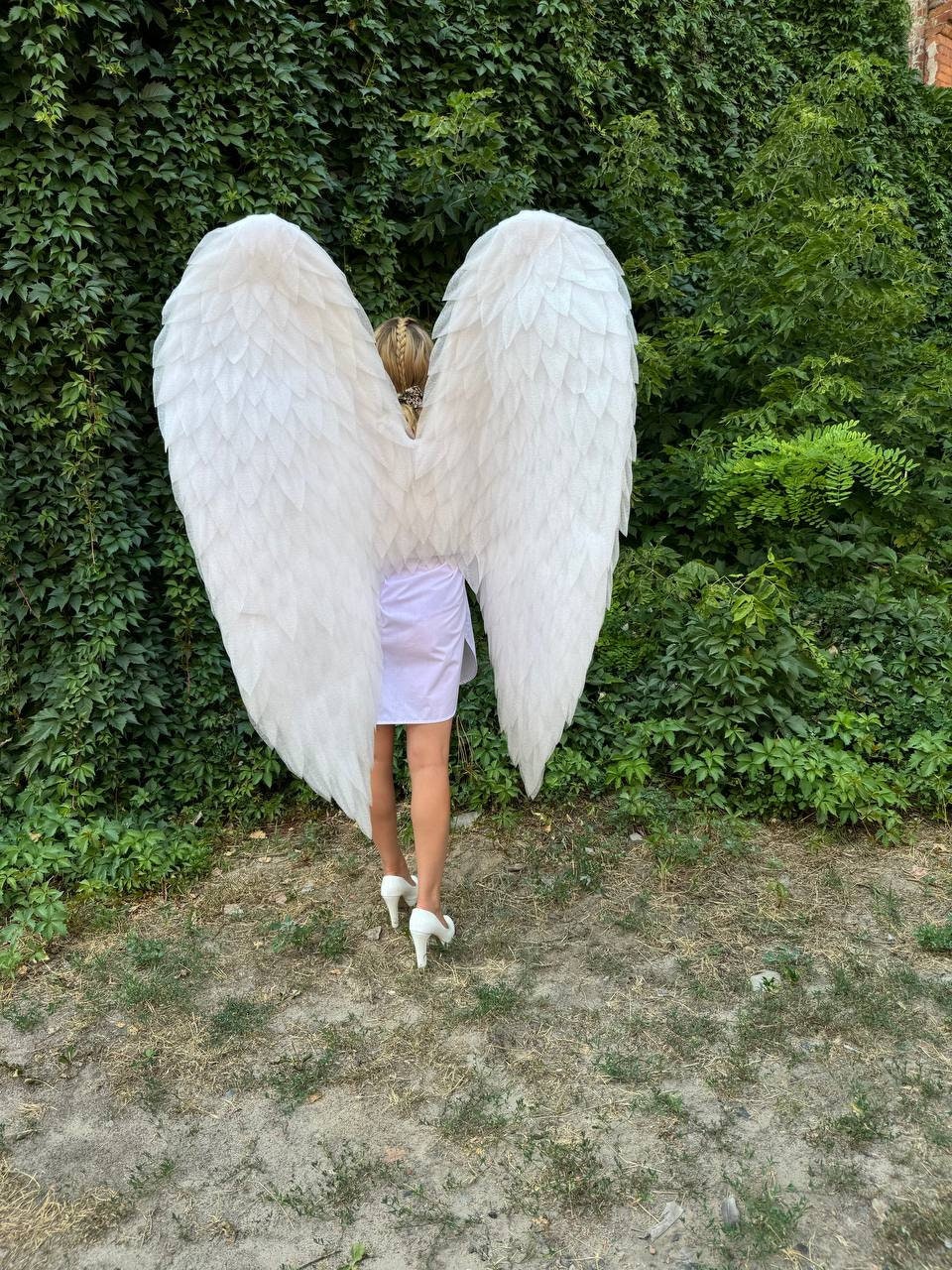May include: Large, white feathered angel wings, shaped like a heart, are the focal point of this image. The wings are worn by a person in a white dress and white heels, standing in front of a lush green wall of foliage. The wings are the main product.